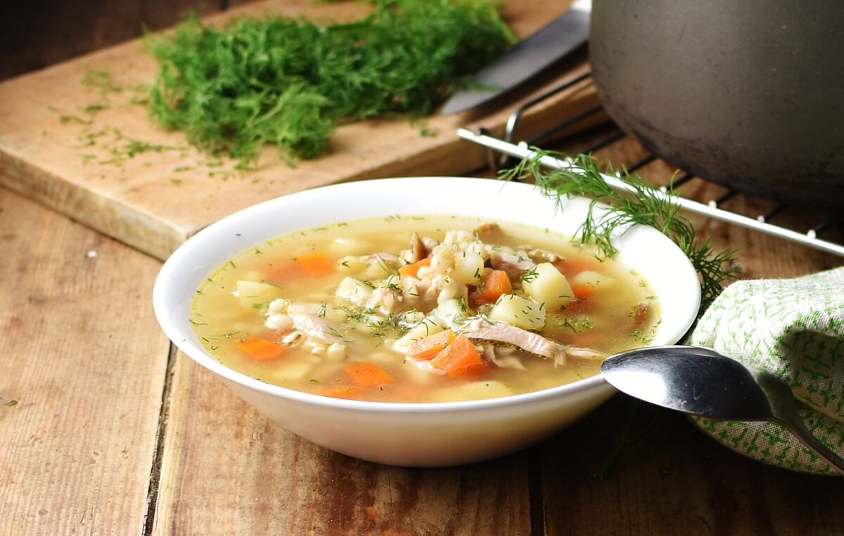 Chunky vegetable krupnik soup in white bowl with spoon, fresh dill and black pot in background.