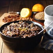 Wheat berries, nuts and raisins mixture with spoon in black bowl, with raisins, nuts, fruit and white bowls stacked in background.