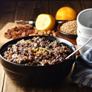 Wheat berries, nuts and raisins mixture with spoon in black bowl, with raisins, nuts, fruit and white bowls stacked in background.