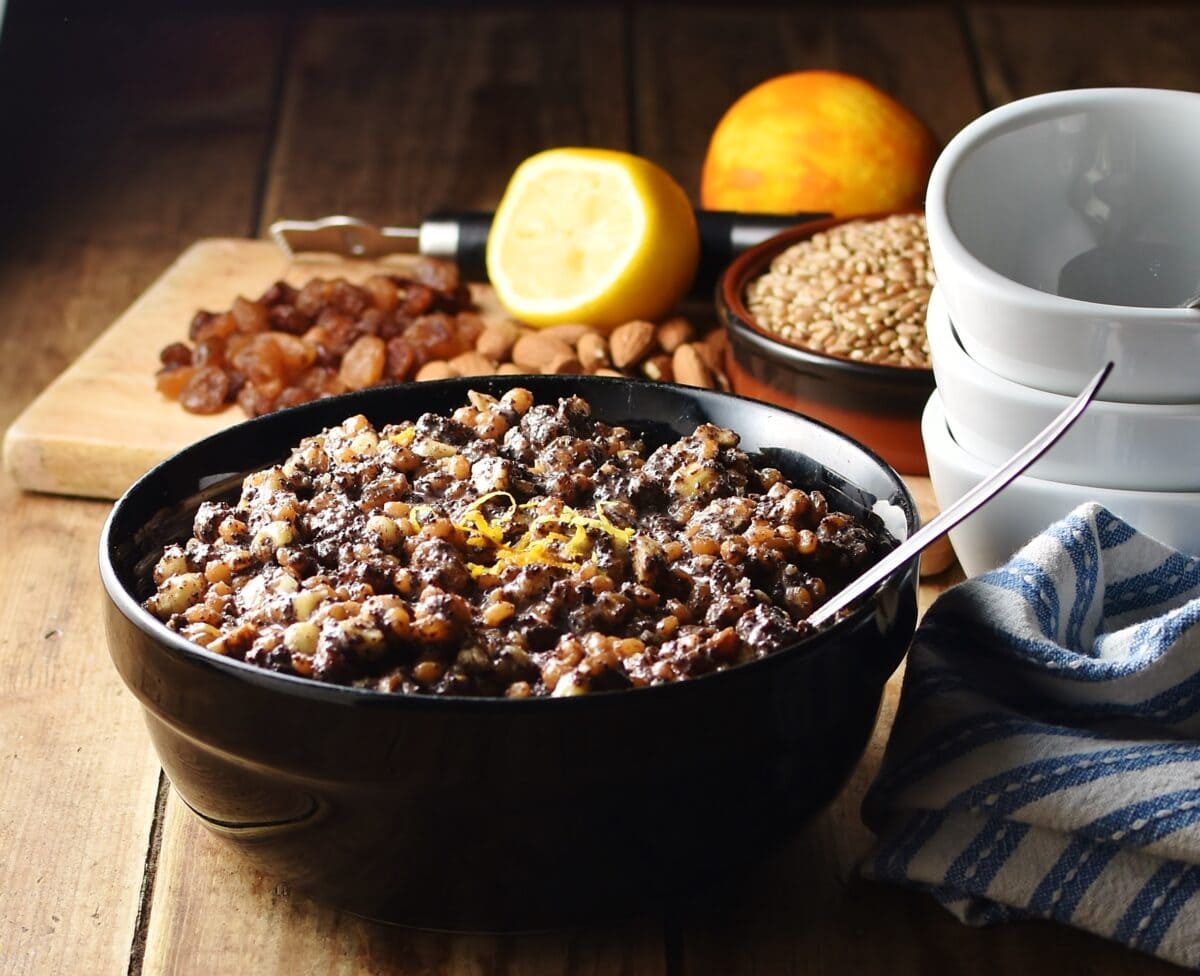 Wheat berries, nuts and raisins mixture with spoon in black bowl, with raisins, nuts, fruit and white bowls stacked in background.