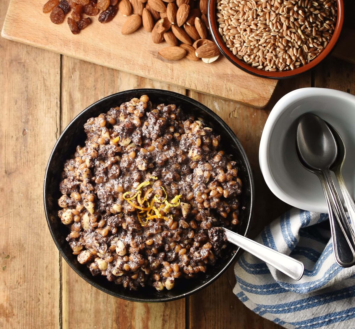 Fruit, wheat and nut kutia mixture in black bowl with spoon, blue-and-white stripy cloth to the right, white dish with spoons and grains and nuts in background.
