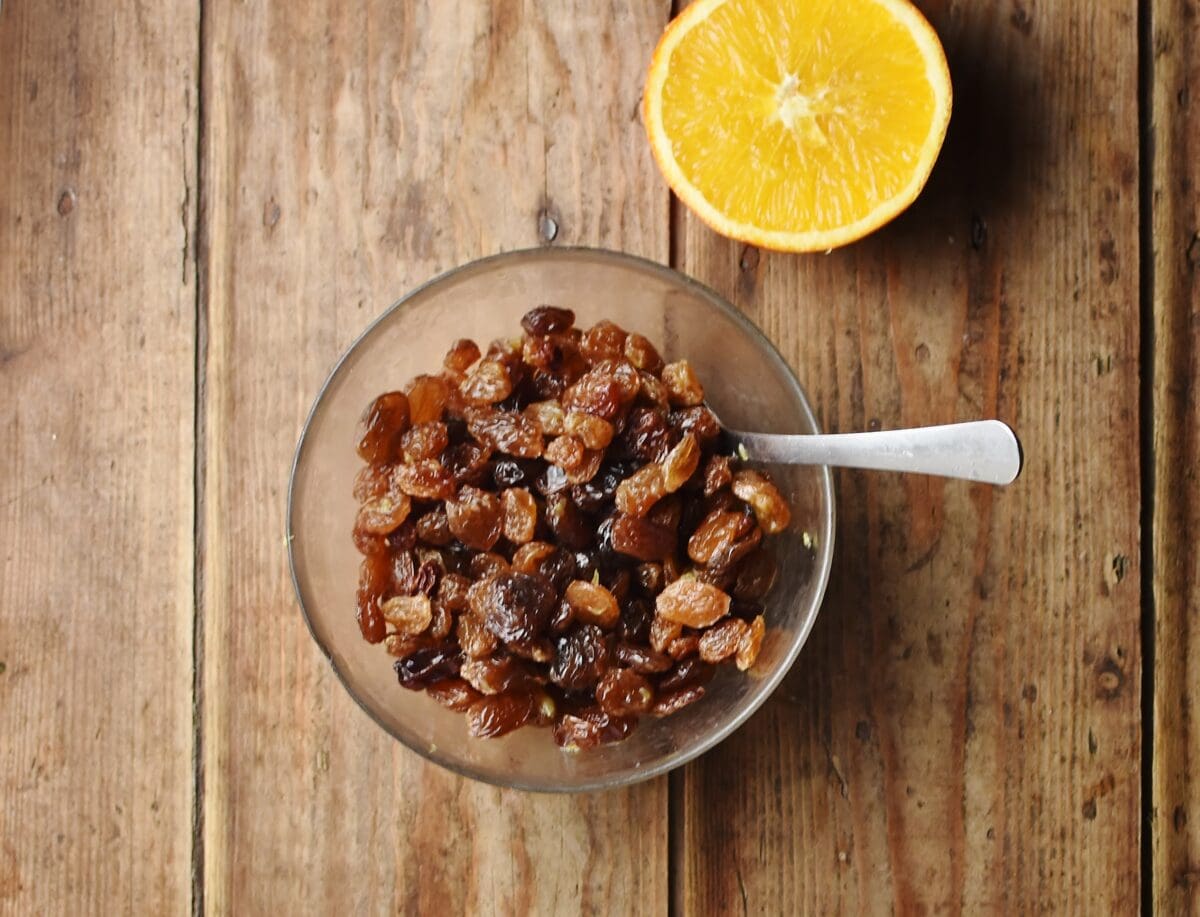 Raisins in bowl with spoon and orange half in background.