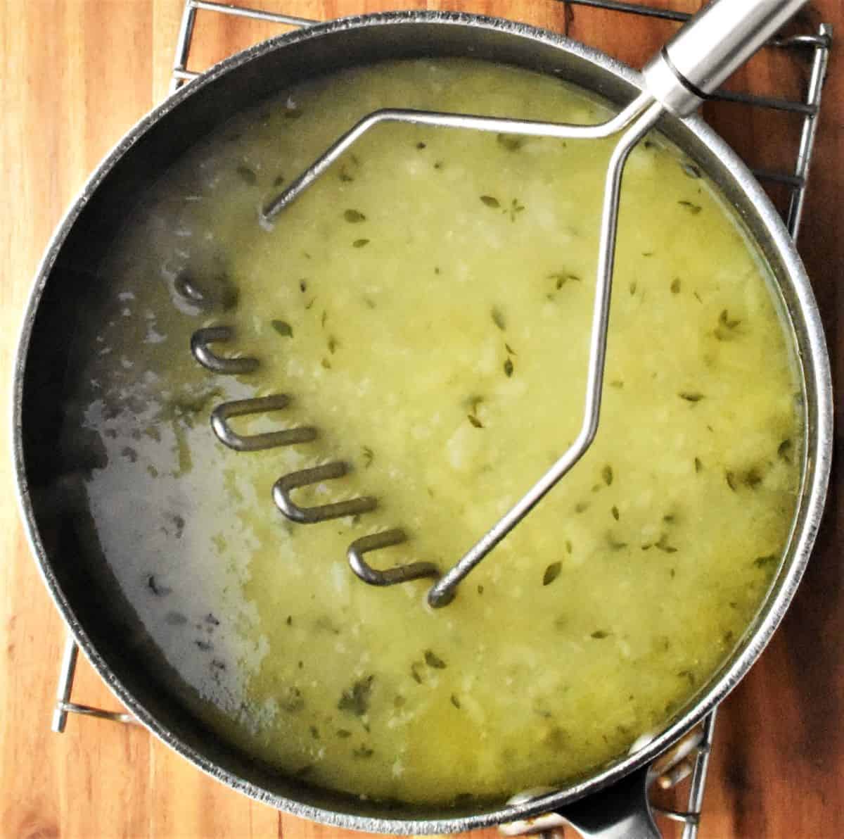 Creamy soup with potato masher in pot.