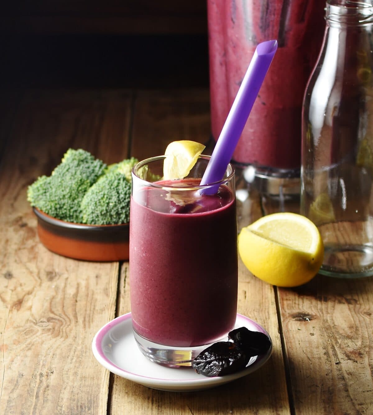 Berry broccoli smoothie in glass with purple straw on top of white saucer with prunes, lemon, broccoli and smoothie in blender in background.