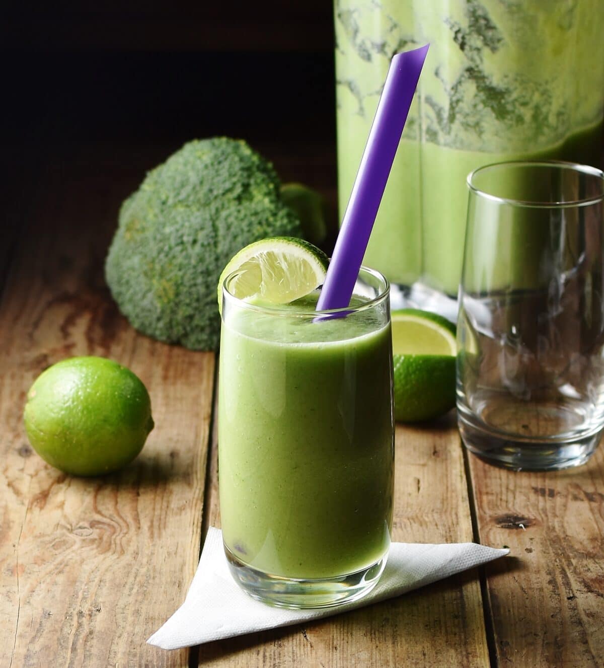Side view of green broccoli smoothie in glass with purple straw on top of folded napkin, with lime, broccoli, smoothie in blender and empty glass in background.