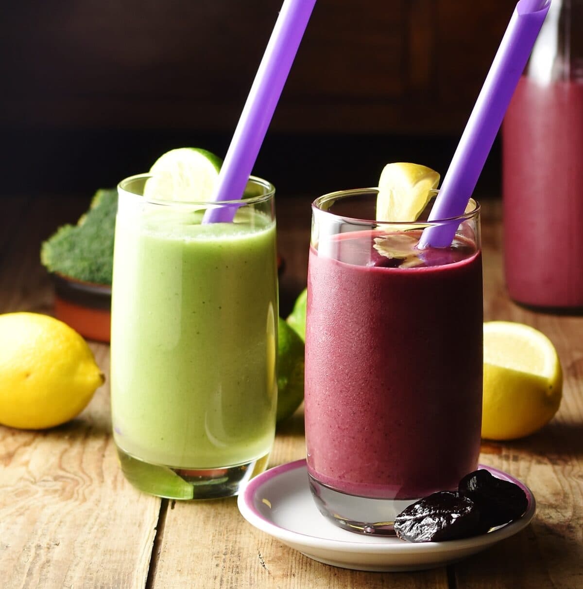 Side view of purple and green smoothies in 2 glasses with purple straws, with lemons, broccoli and smoothie in bottle in background.