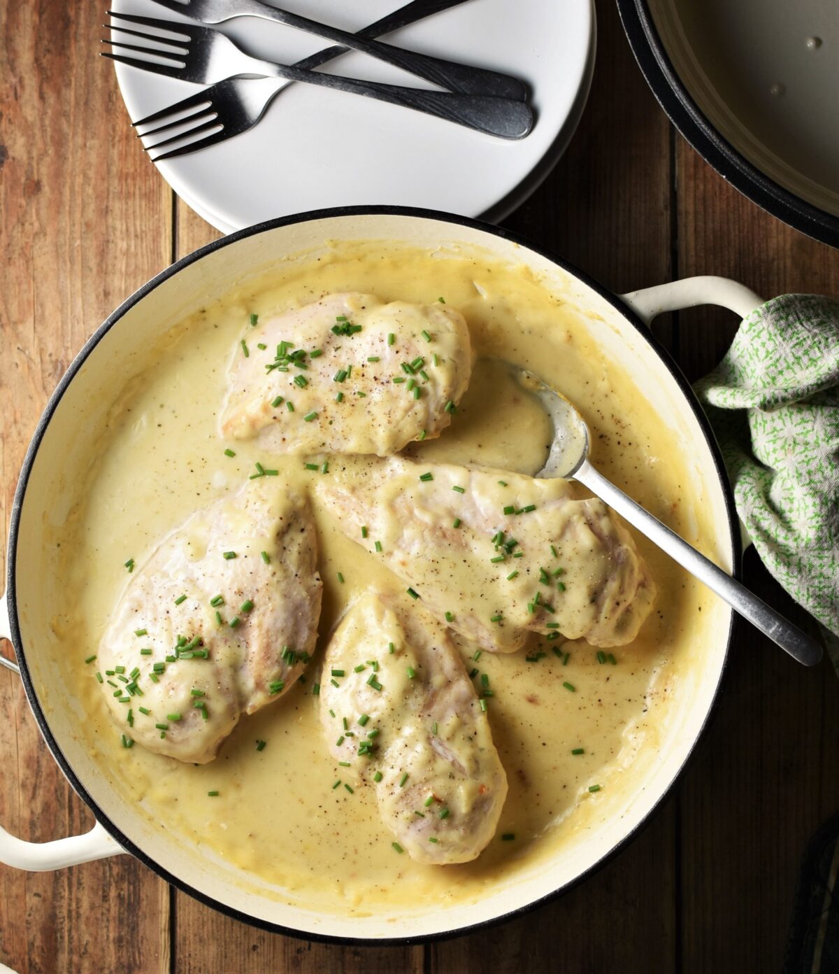 4 chicken breasts with chopped chives in large shallow white pan with spoon, green cloth to the right and plates with forks at the top.