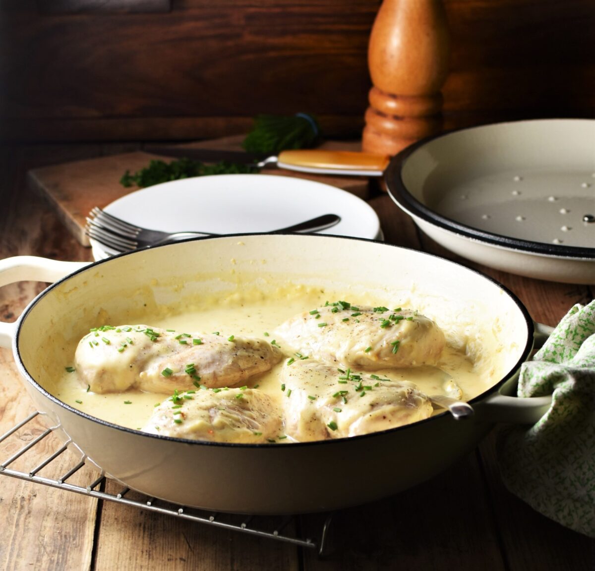 Side view of chicken in white sauce in large shallow white pan, with plates, lid and pepper grinder in background.