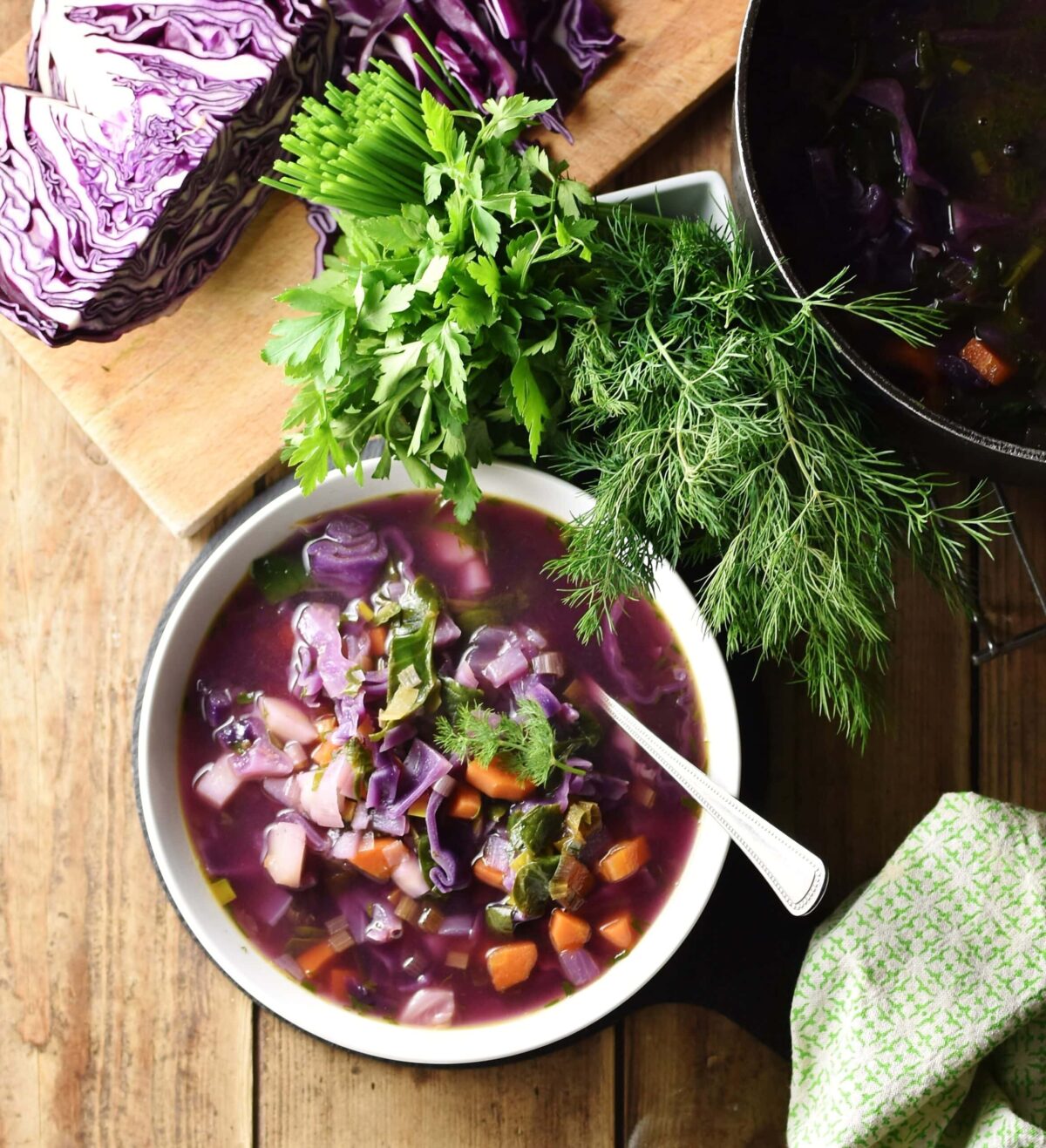 Chunky vegetable red cabbage soup in white bowl with spoon, green cloth, fresh herbs, red cabbage and pot in background.