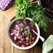 Top down view of red cabbage vegetable soup in white bowl with spoon, dill, parsley, green cloth and red cabbage in background.