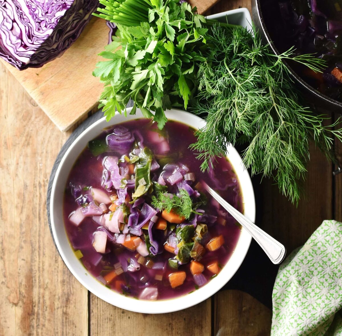 Chunky red cabbage vegetable soup in white bowl with spoon, dill, parsley, green cloth and red cabbage in background.