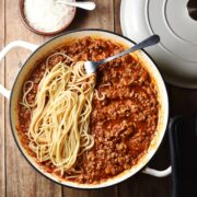 Top down view of turkey bolognese sauce with spaghetti and fork in large white shallow dish, with grated cheese in small dish and white lid in background.