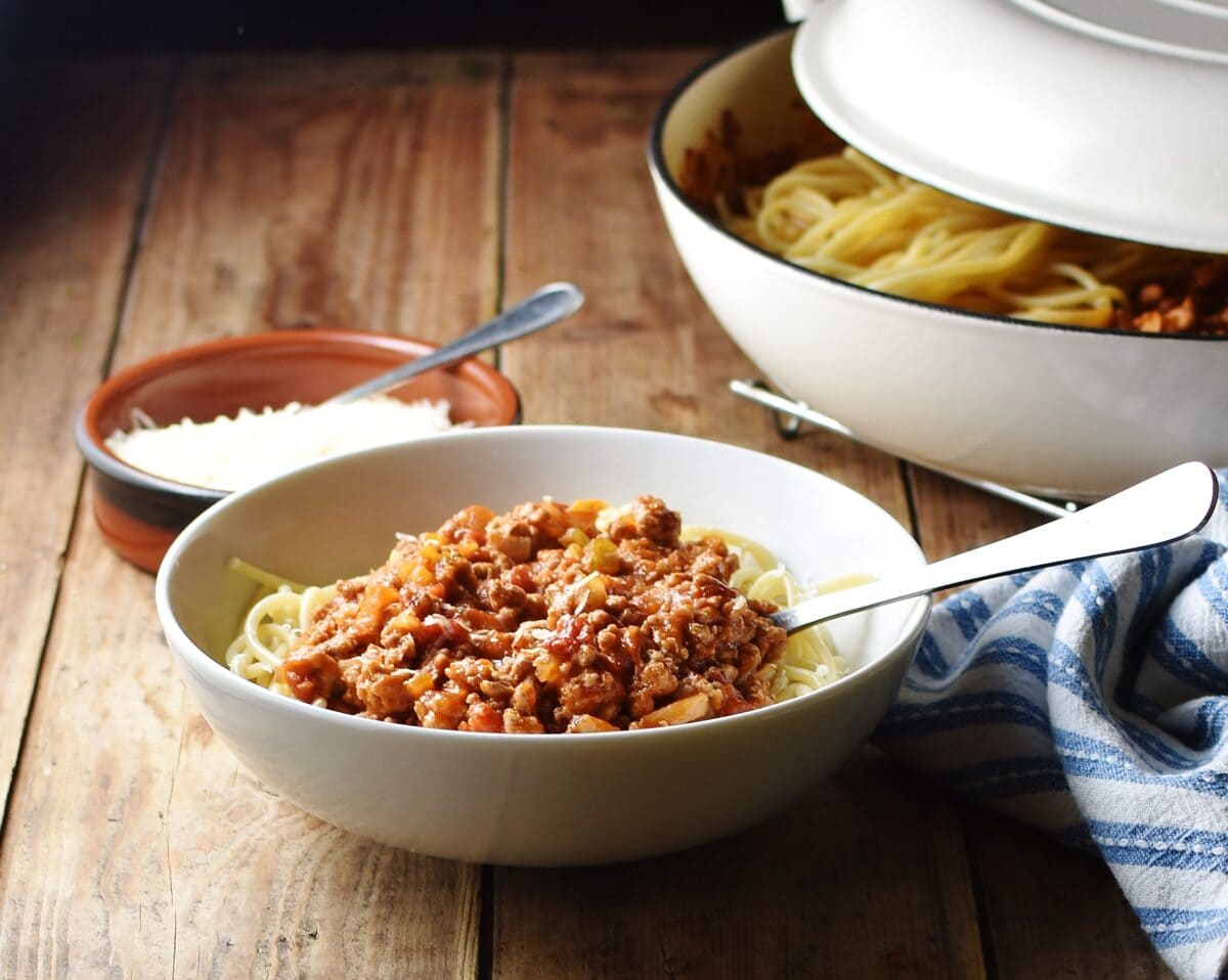 Side view of turkey spaghetti sauce in white bowl with fork, grated cheese in brown dish with spoon, white pan with lid and stripy blue cloth in background.