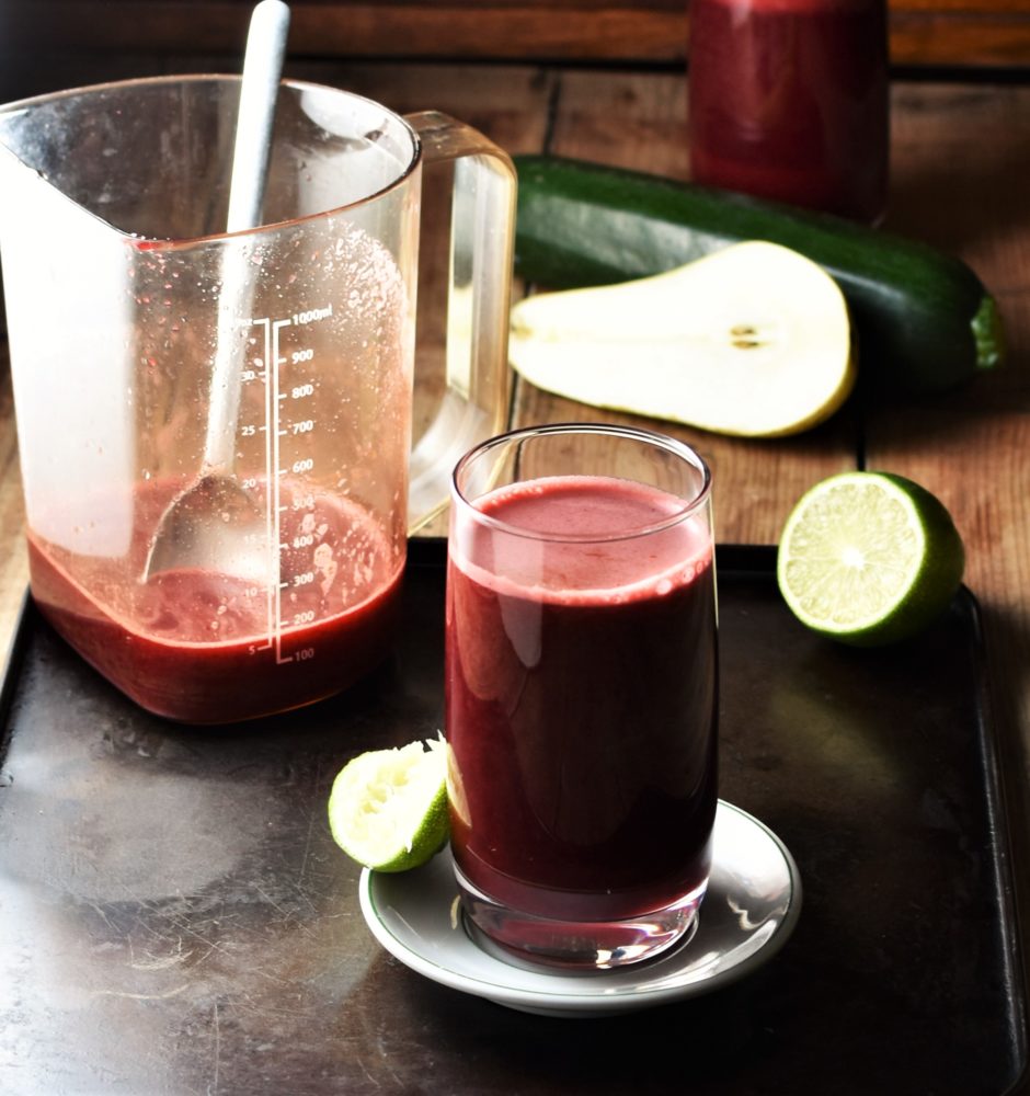 Side view of beet and carrot juice in glass on top of white saucer, with pear, lime, zucchini and juice in container with spoon in background.