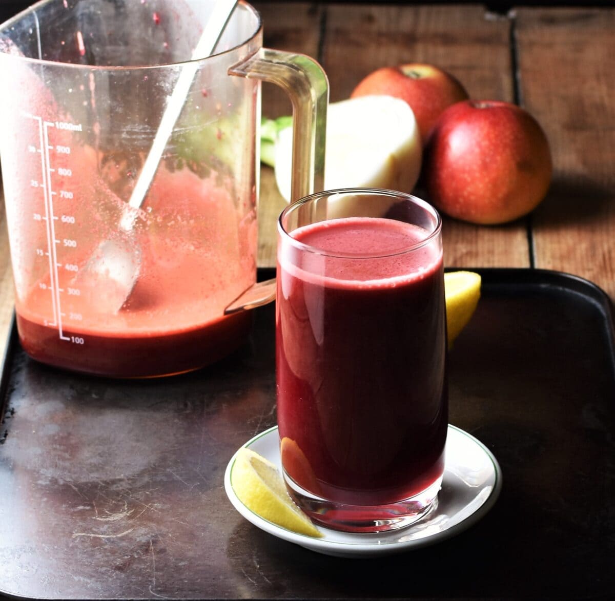 Side view of beet juice in glass on top of white saucer, with apples, fennel and juice in container with spoon in background.