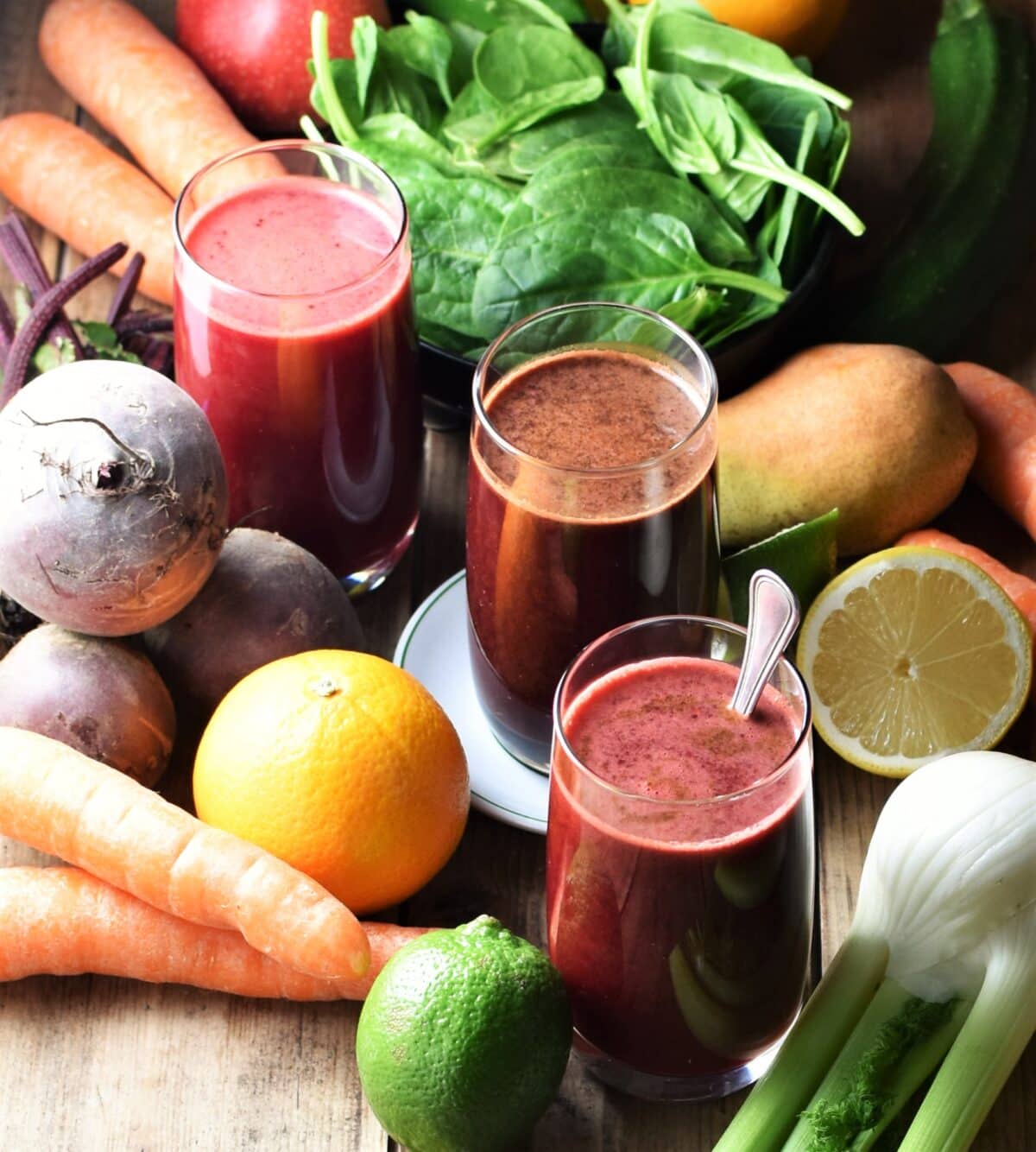 3 glasses with dark juice surrounded by fresh fruits and vegetables.