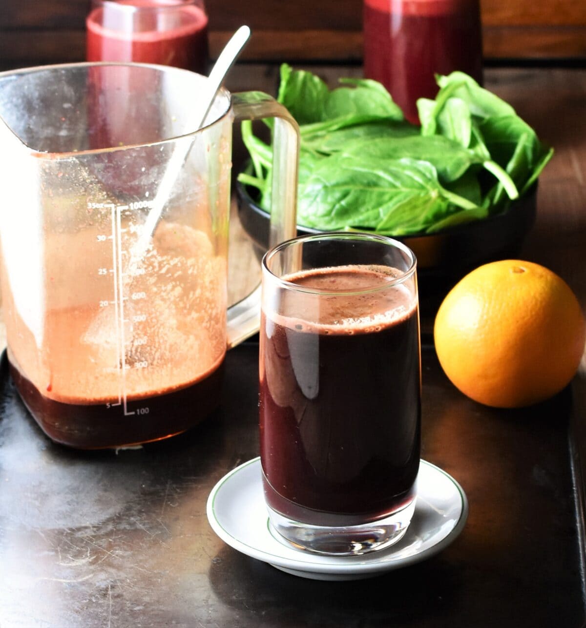 Side view of beet spinach juice in glass on top of white saucer, with orange, spinach and juice in measuring container with spoon in background.