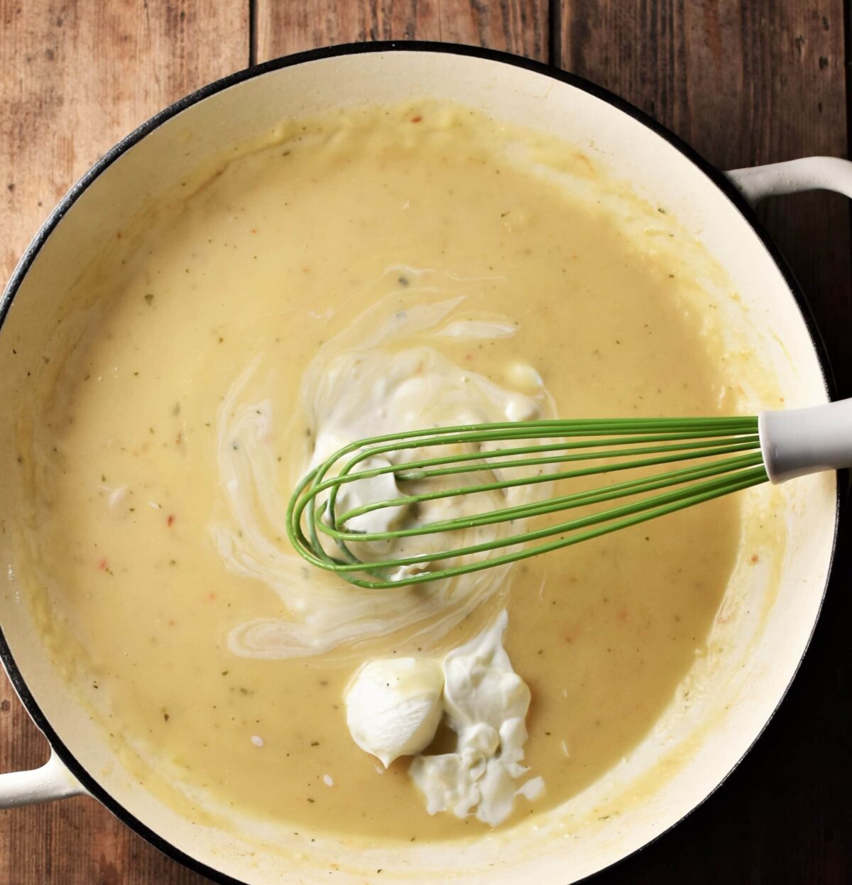 Creamy sauce with dollop of yogurt and green whisk in large white pan.