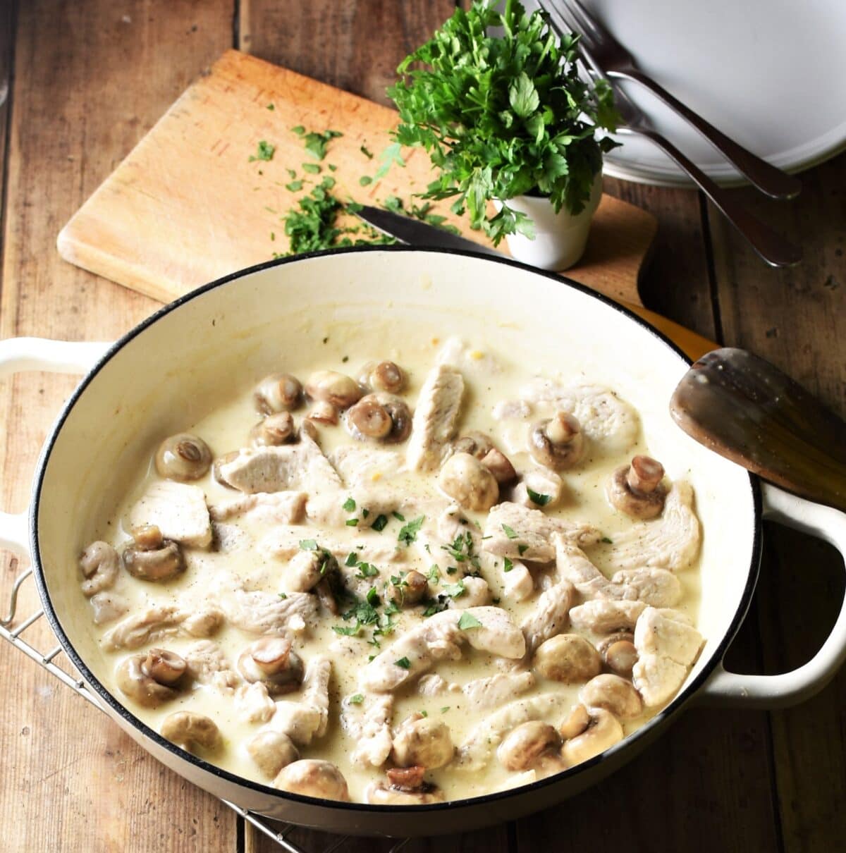 Creamy turkey mushroom stroganoff in large white shallow pan with herbs on top of wooden board and plates in background.