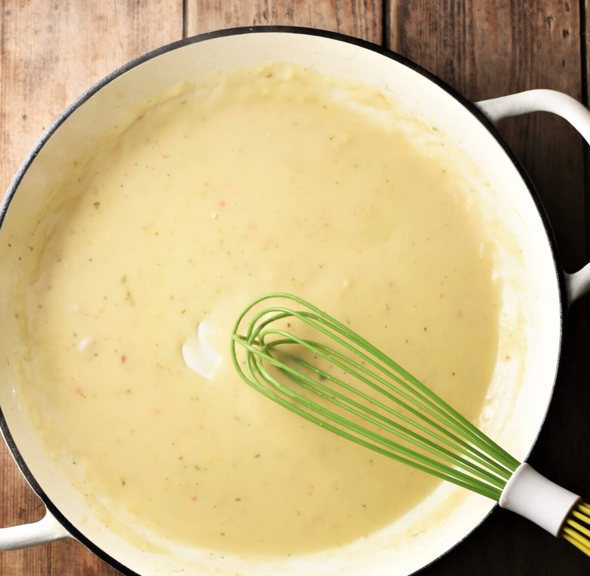 Creamy sauce in large white pan with green whisk.