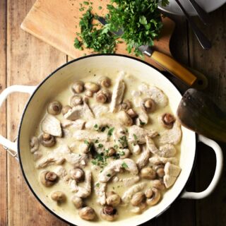 Creamy turkey and mushroom in large white pan, with chopped herbs on top of wooden board with knife in background.