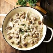 Creamy turkey and mushroom in large white pan, with chopped herbs on top of wooden board with knife in background.