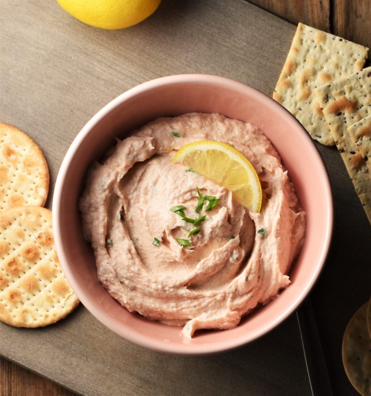 Creamy salmon spread in pink bowl with crackers and lemon on top of grey wooden board.