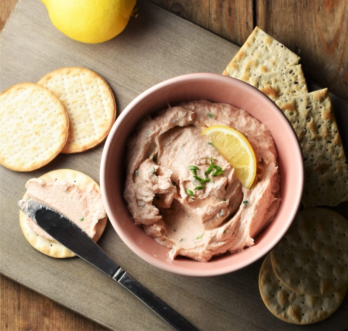 Salmon spread in pink bowl, crackers, knife and lemon on top of grey board.