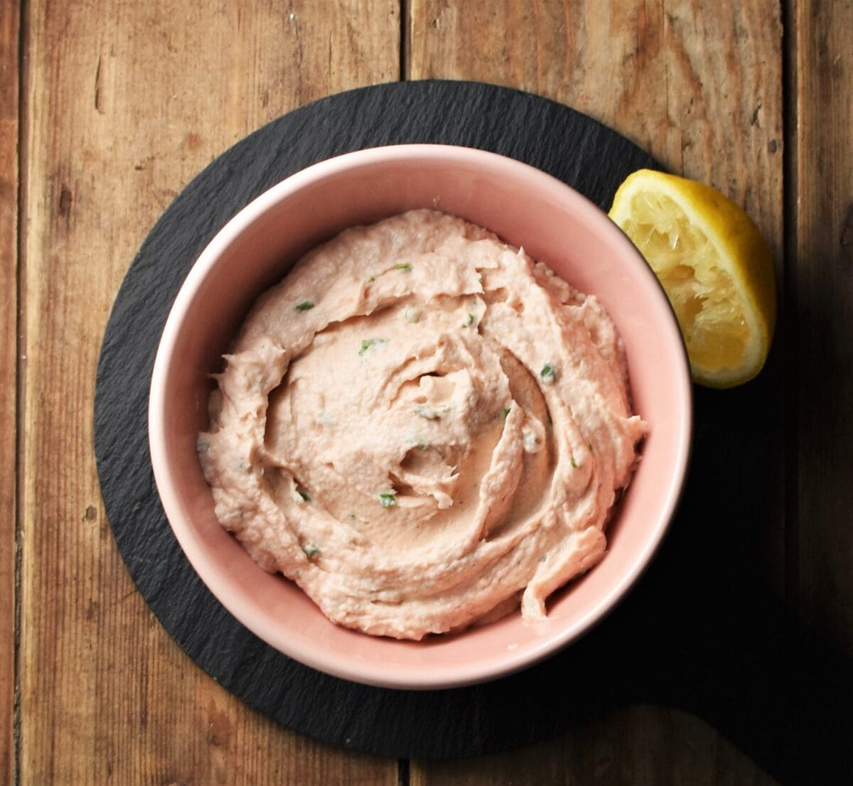 Creamy salmon spread in pink bowl with halved lemon on black platter.