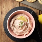 Creamy salmon spread with lemon slice in pink bowl on black plate with crackers and lemon in background.