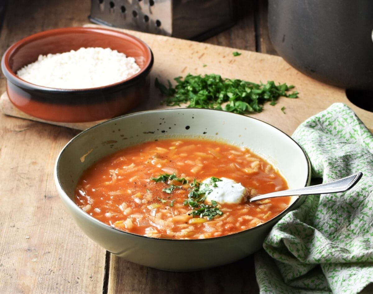 Side view of tomato soup with rice in green bowl with spoon, rice in brown dish, chopped herbs and green cloth in background.