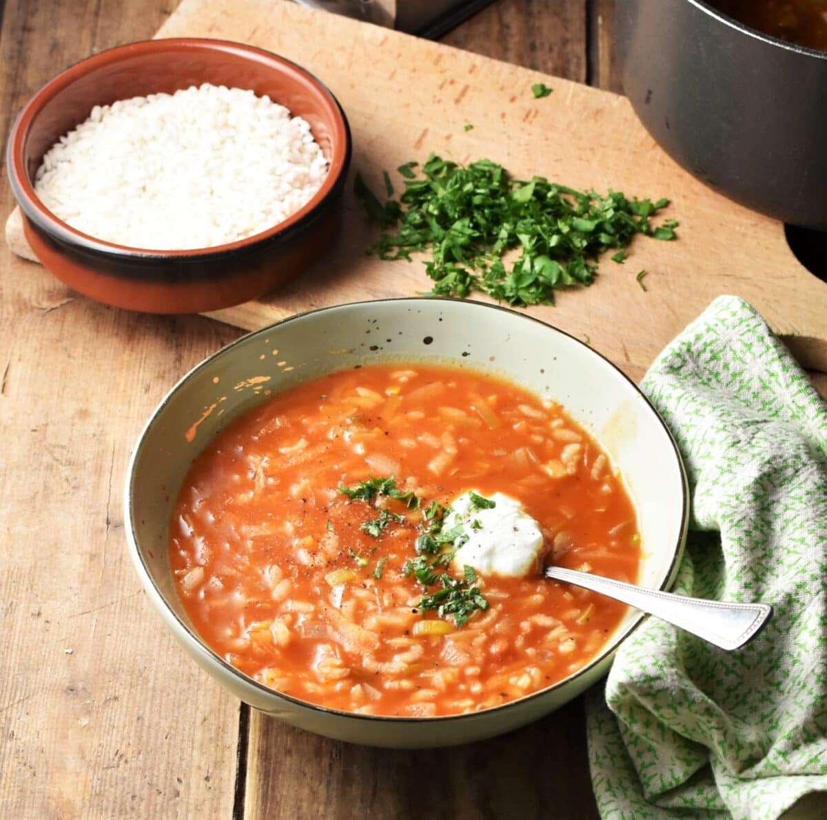 Chunky rice tomato soup with yogurt and herbs in green bowl with spoon, green cloth to the right, rice in brown dish and chopped herbs on wooden board in background.