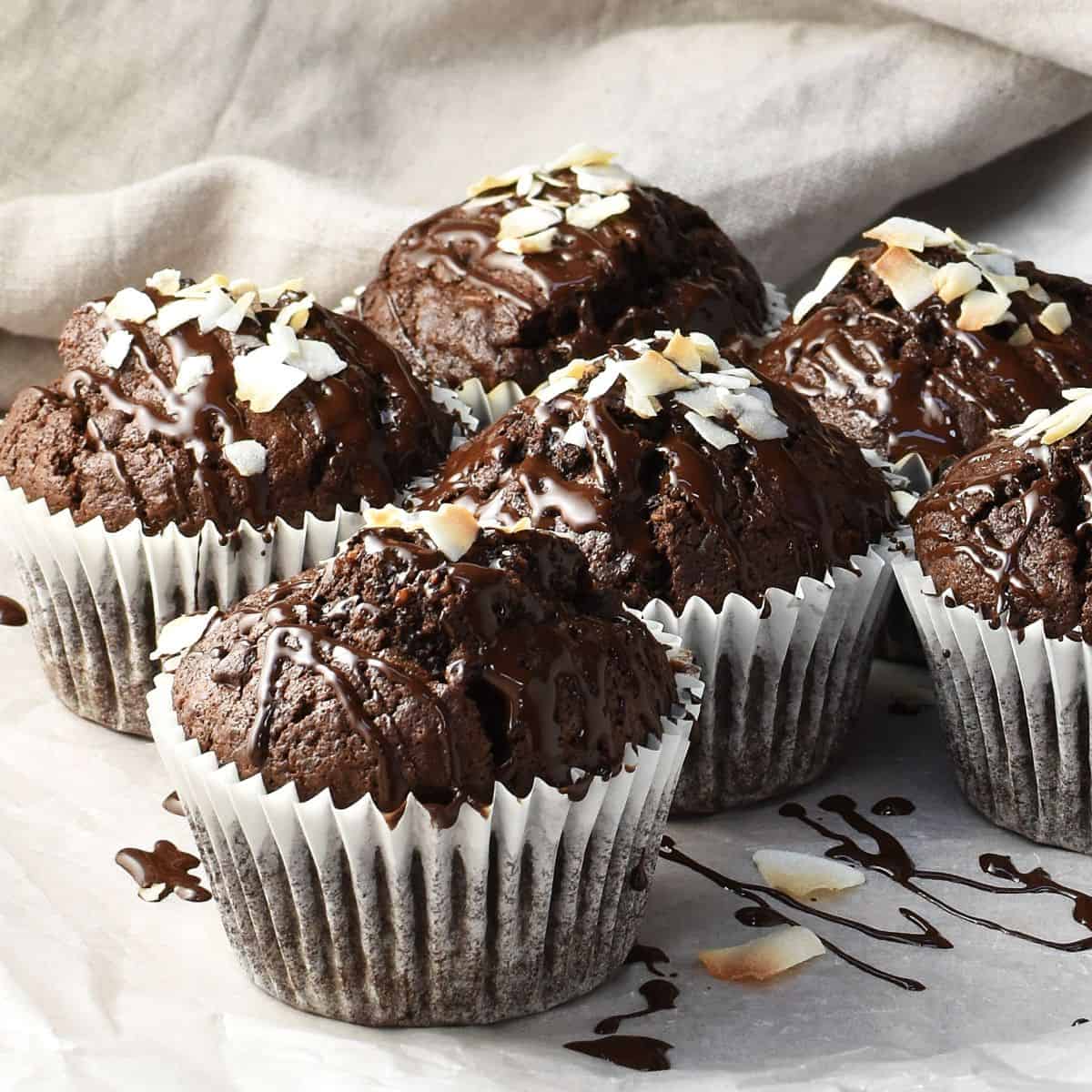Side view of chocolate muffins in white cases with chocolate drizzle and coconut flakes on top.