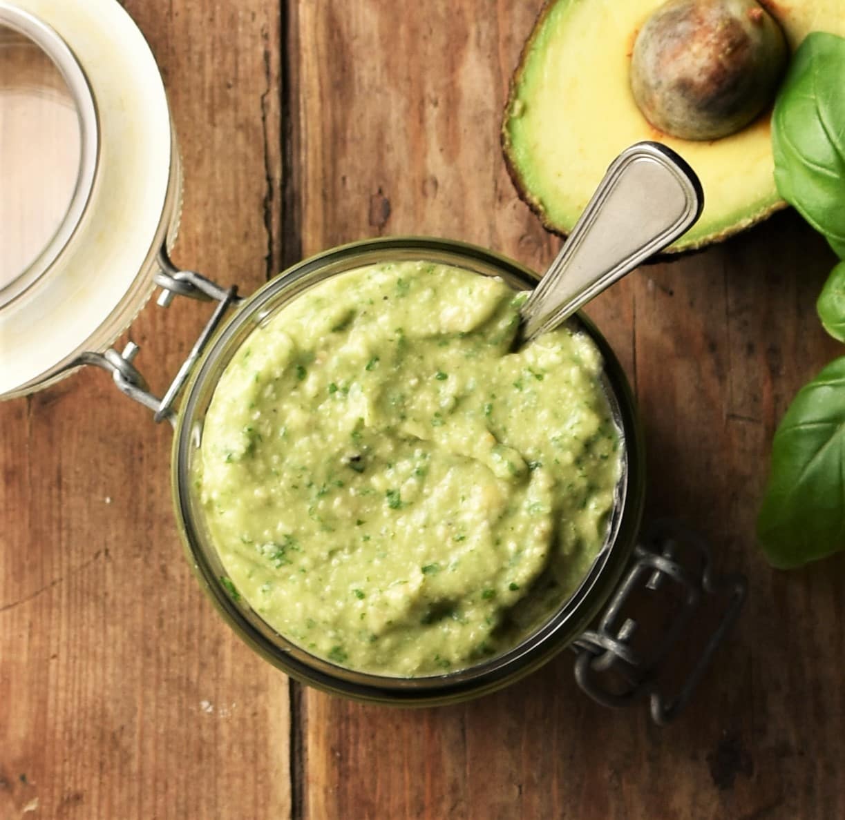 Top down view of avocado pesto in open jar with spoon and avocado in background.