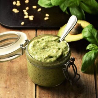 Side view of avocado pesto in jar with spoon, avocado, basil and pine nuts in background.
