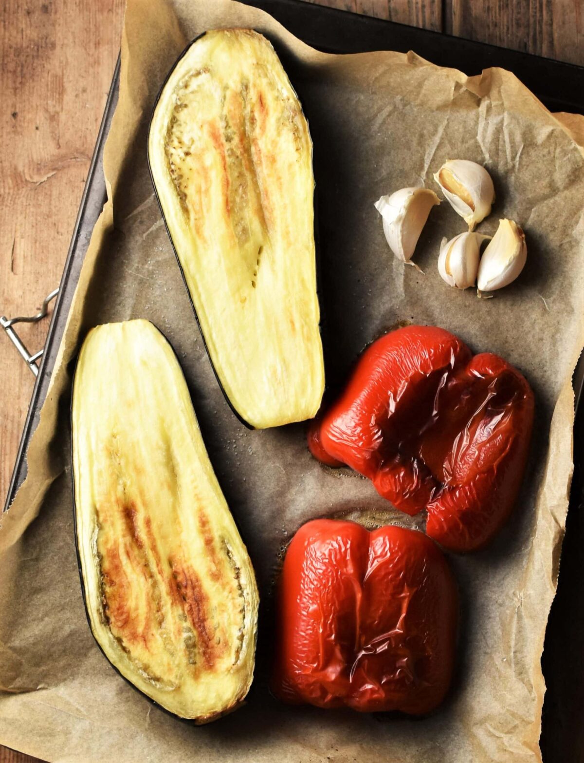 Top down view of roasted eggplant and red pepper with 4 garlic cloves on parchment.