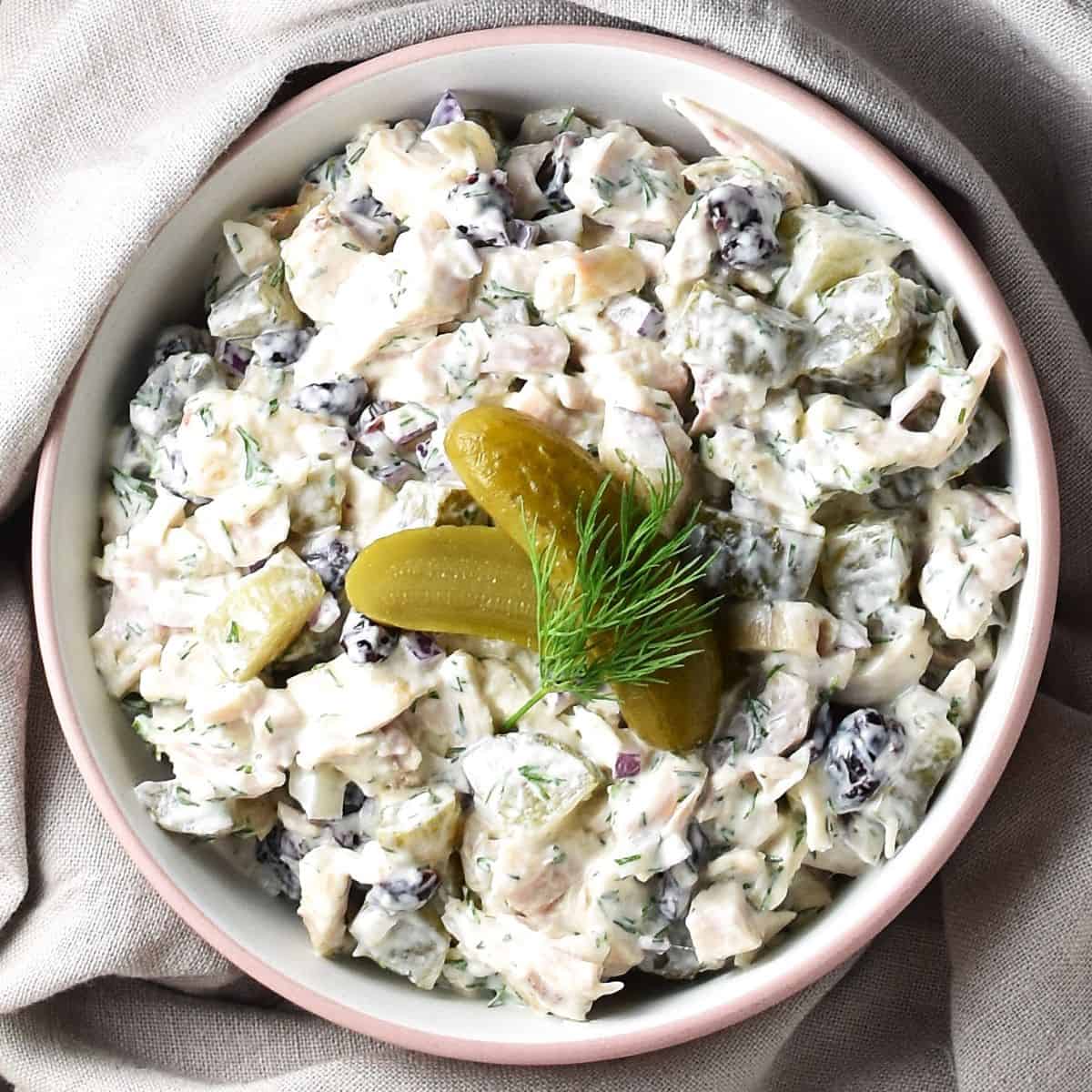 Top down view of chicken salad with dill pickles in white bowl with beige cloth in background.