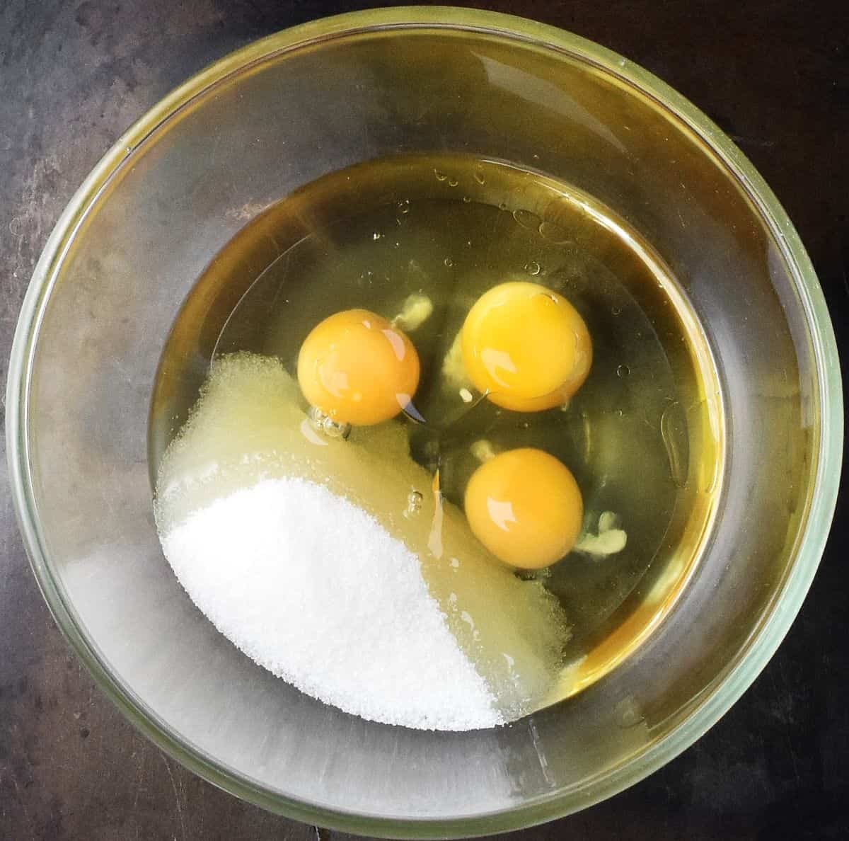 Top down view of 3 raw eggs, oil and sugar in glass bowl.