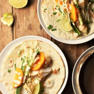 2 open fish tacos with vegetables on white plates with lime in background.