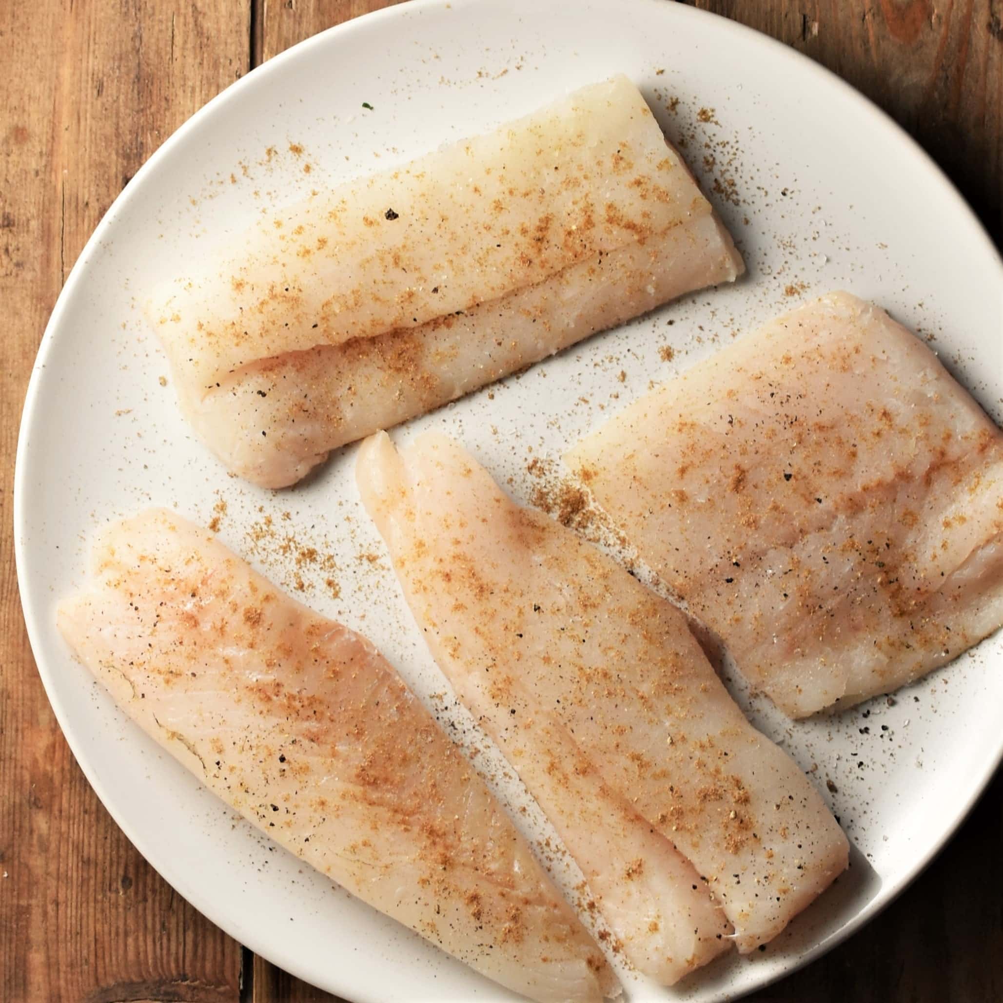 4 large fish fillets with seasoning on white plate.