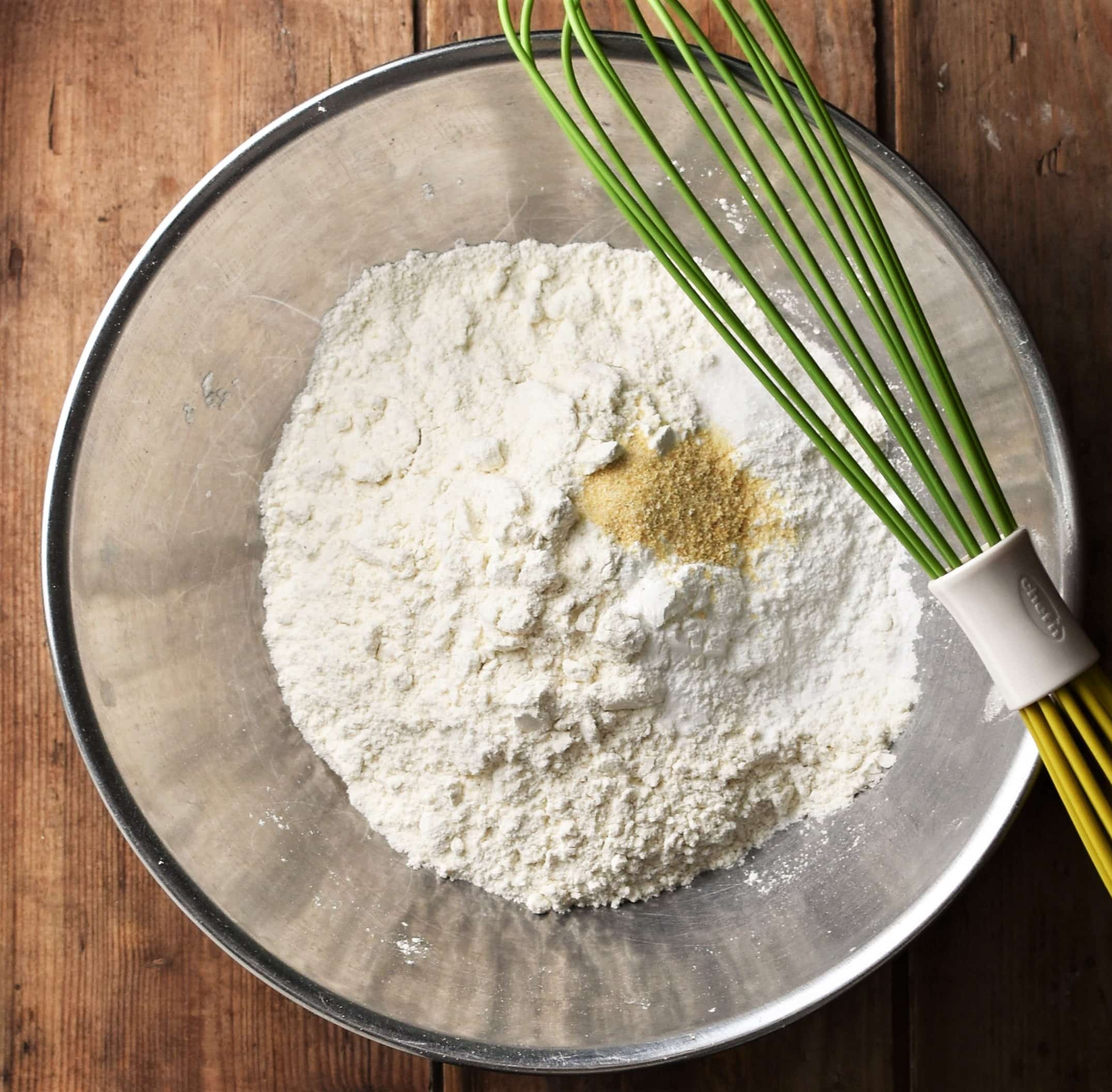 Flour and spice in large metal bowl with green whisk on top.