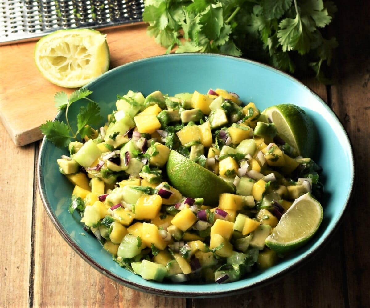 Chunky mango avocado salsa with lime wedges in large blue bowl with lime, zester and cilantro in background.