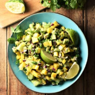 Mango, avocado and cucumber salsa with lime wedges in blue bowl with lime and cilantro in background.