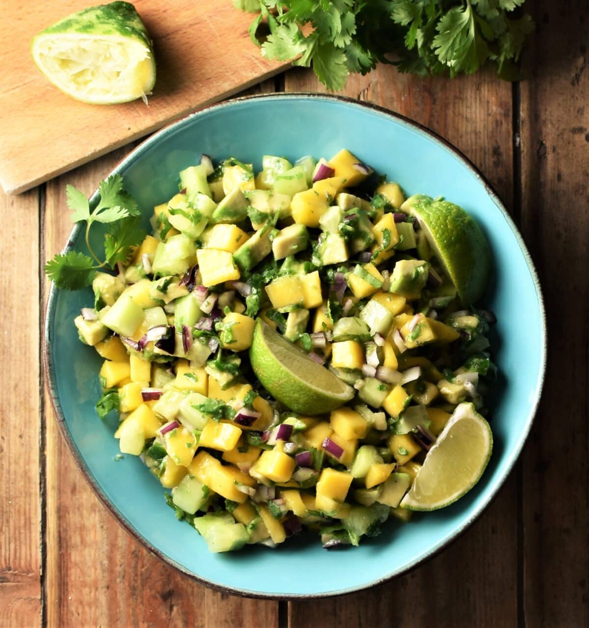 Mango, avocado and cucumber salsa with lime wedges in blue bowl with lime and cilantro in background.