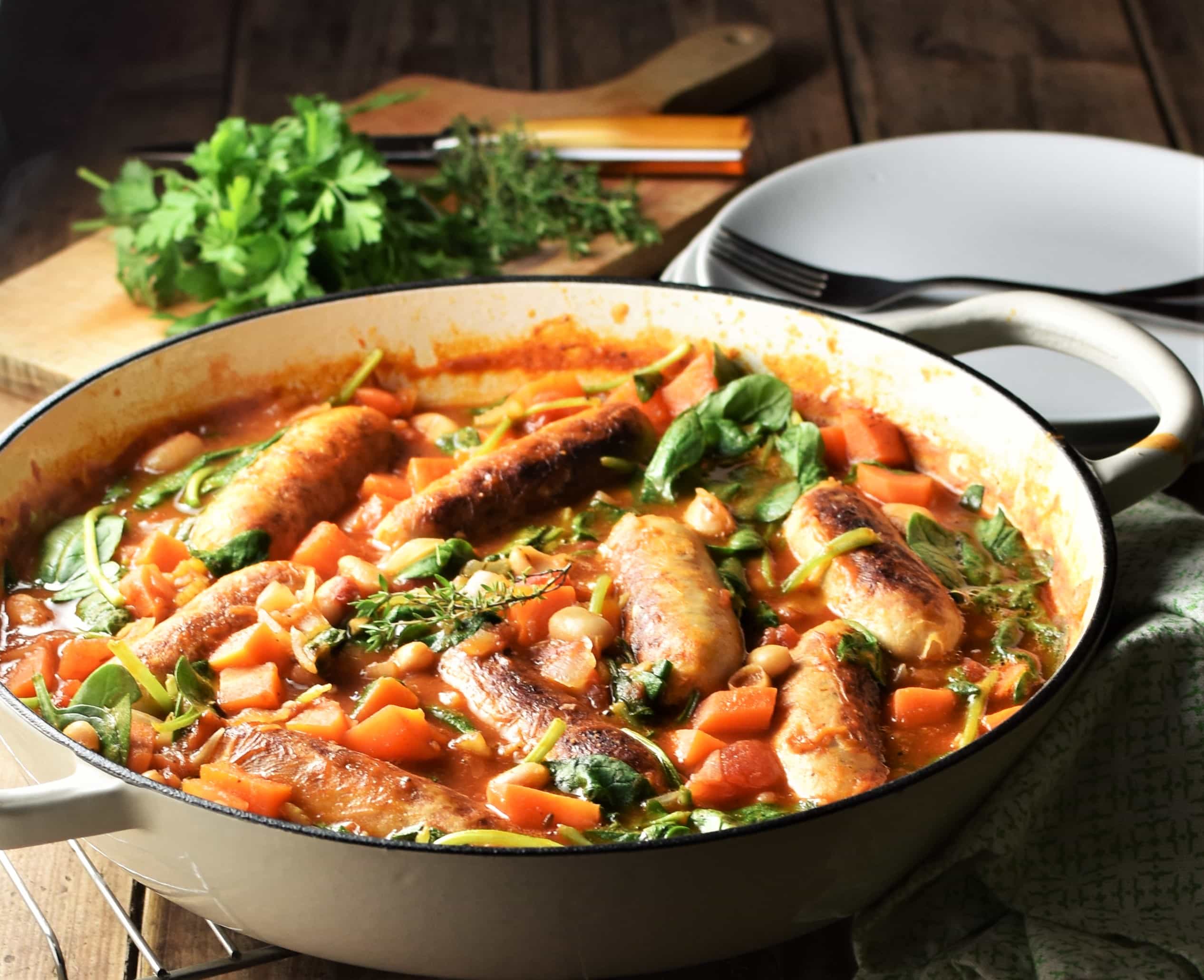 Side view of bean sausage casserole with spinach in white pan, with herbs and plates in background.