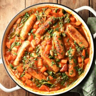 Sausage and bean casserole with vegetables and herbs in large white pan.