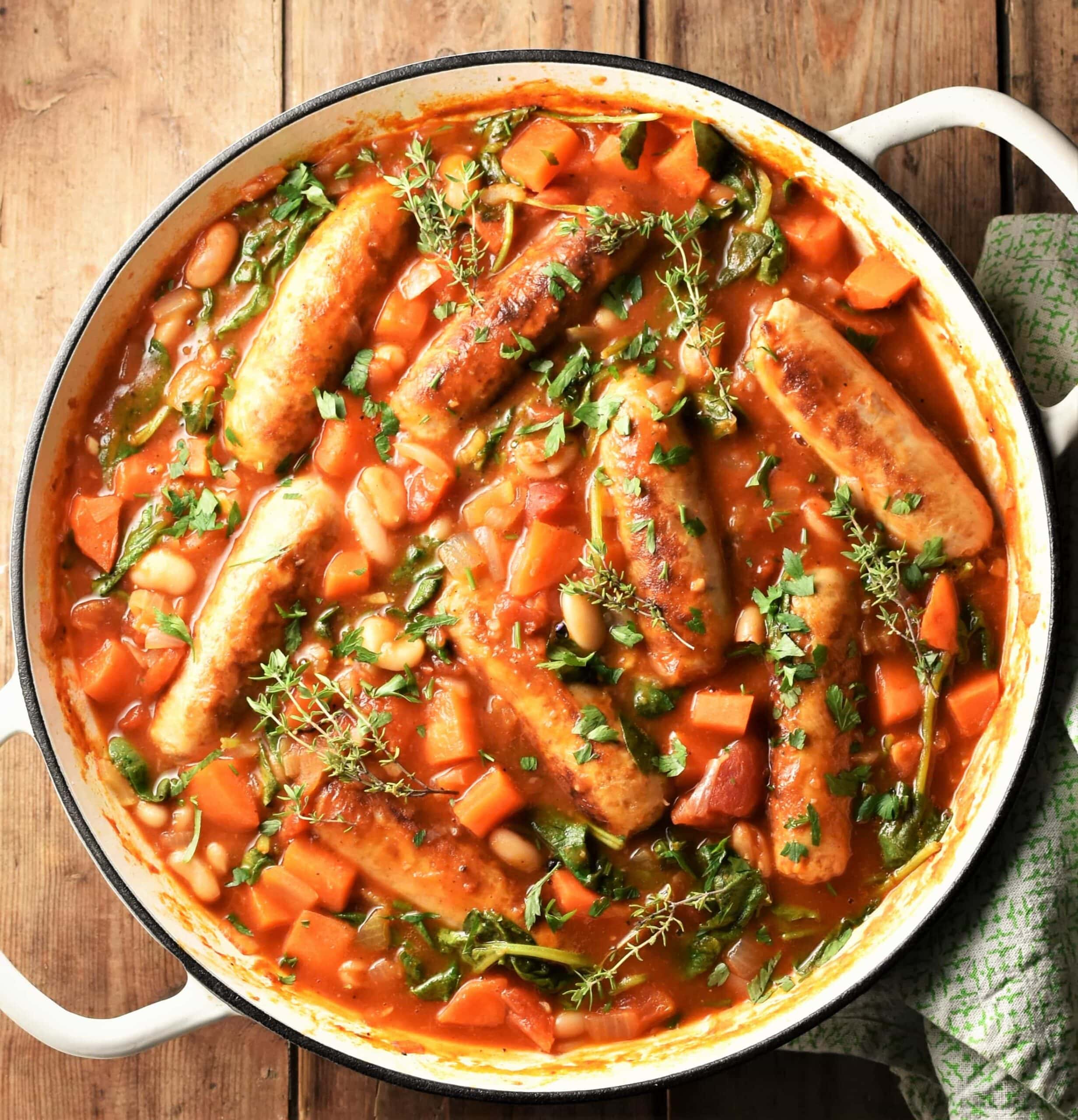 Sausage and bean casserole with vegetables and herbs in large white pan.