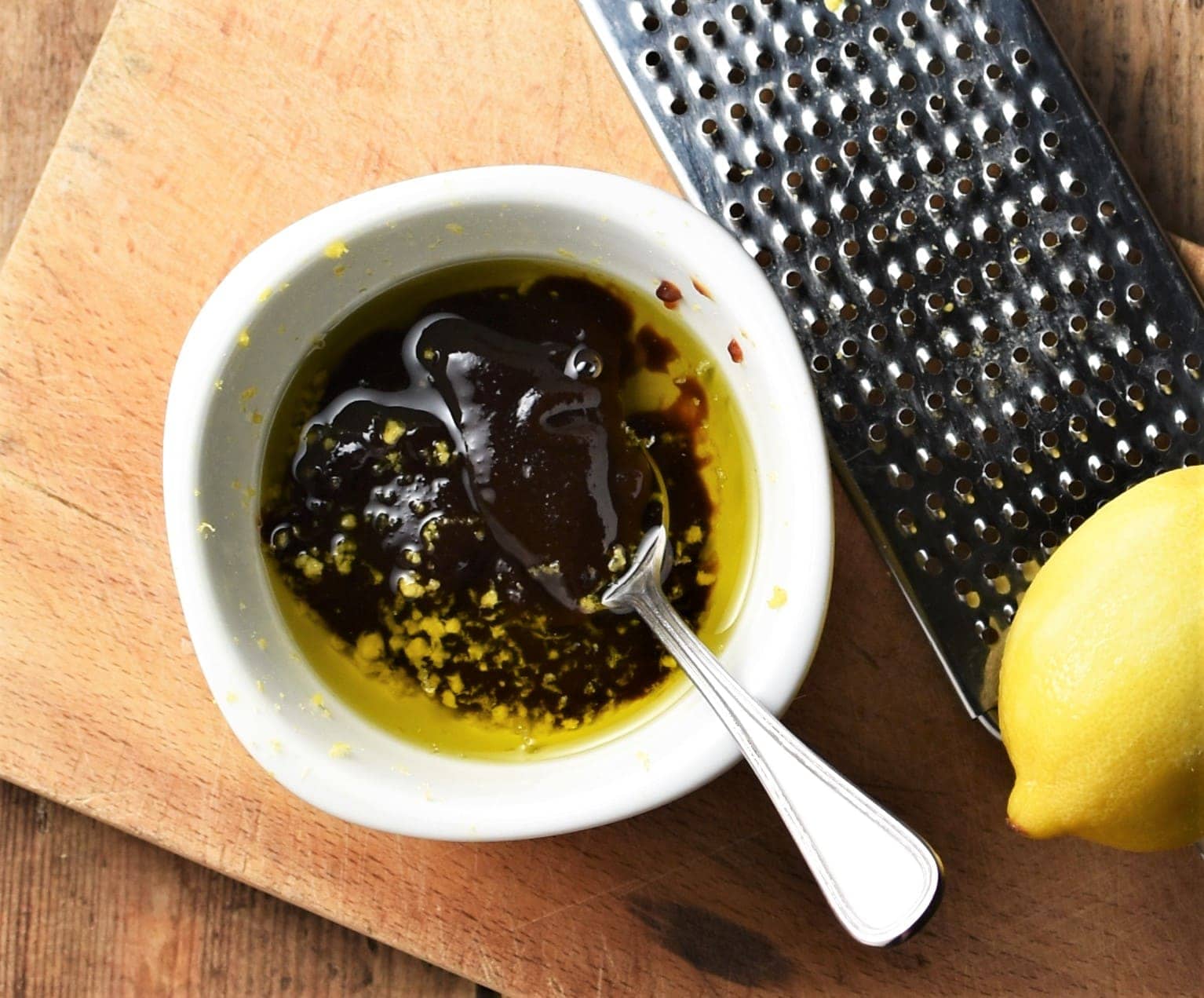 Hoisin and oil in white bowl with spoon, lemon and zester on top of wooden board.