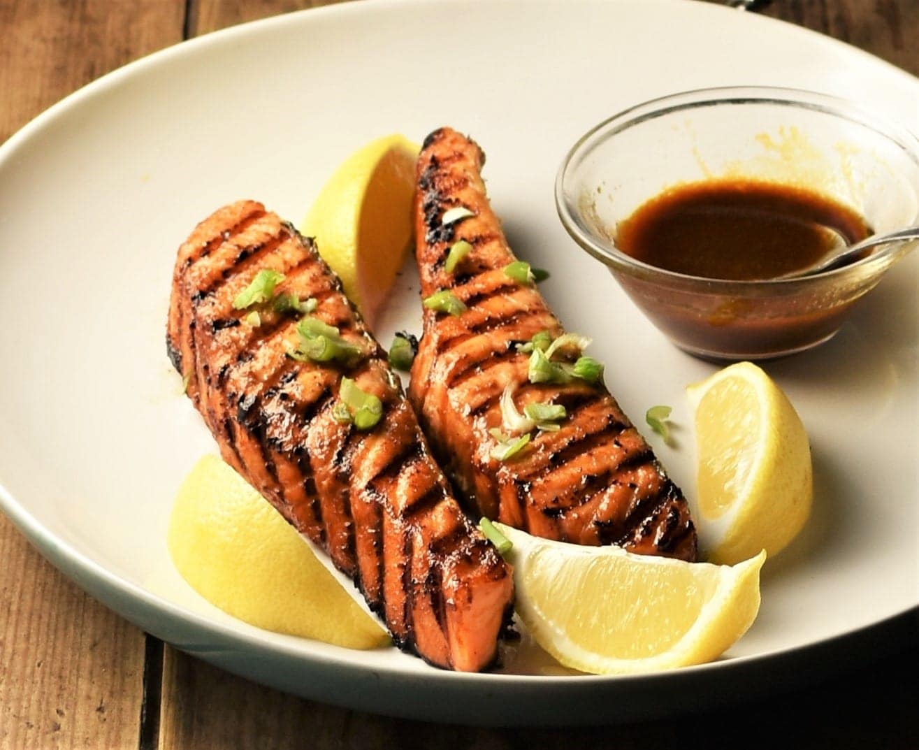 2 grilled salmon fillets with lemon wedges and sauce in small dish.