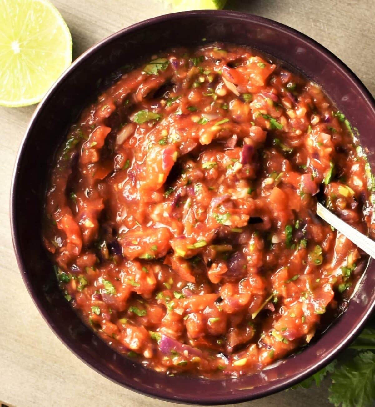 Top down view of tomato salsa in bowl with spoon, limes and cilantro in background.